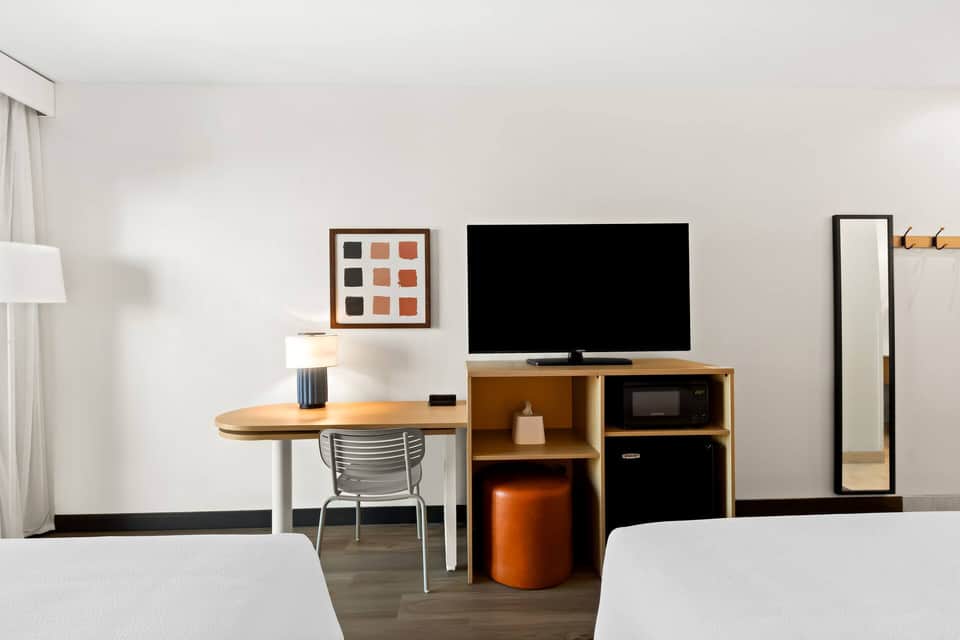 Modern hotel room with desk, TV stand, queen bed, and warm wood furnishings