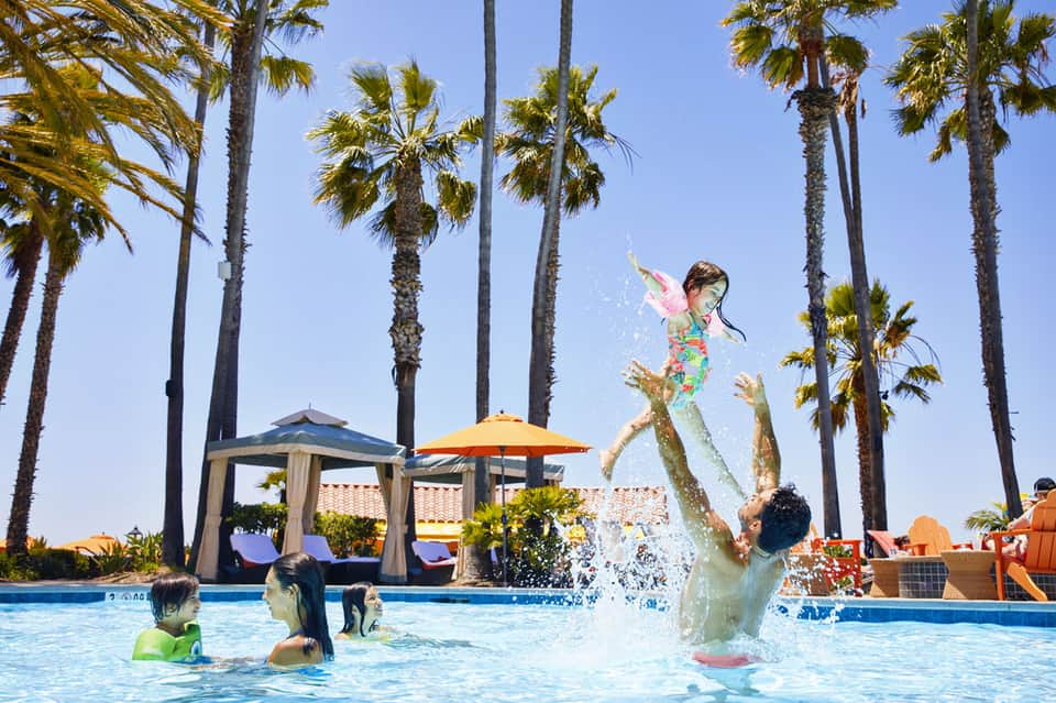 Family enjoying resort pool with children playing in shallow water under palm trees on sunny day