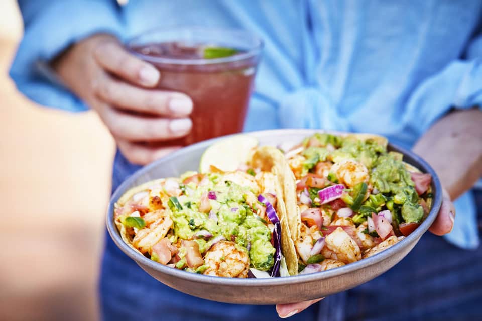Person in blue shirt holding plate of shrimp tacos with guacamole and red beverage