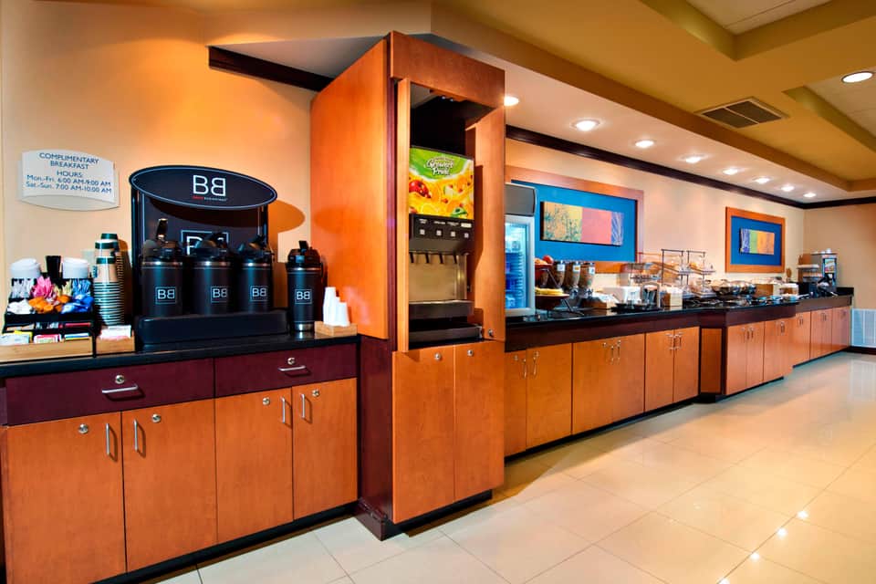 Hotel breakfast area with orange wood cabinetry, coffee station, and beverage dispensers