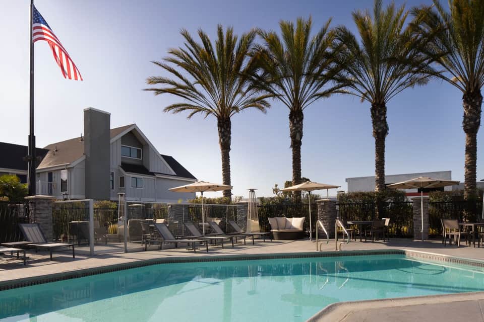 Coastal pool area with palm trees, umbrella shade, lounge chairs, and modern cottage-style building