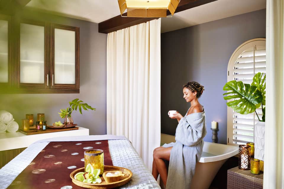 Woman in spa robe holding coffee cup beside luxurious soaking tub with tropical plants