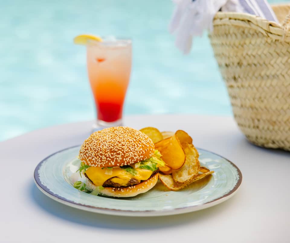 Beachside burger meal with golden fries, colorful drink, and straw beach bag