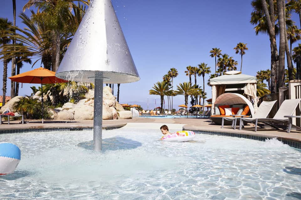 Child in colorful float in shallow pool with cone-shaped fountain and palm trees surrounding resort area