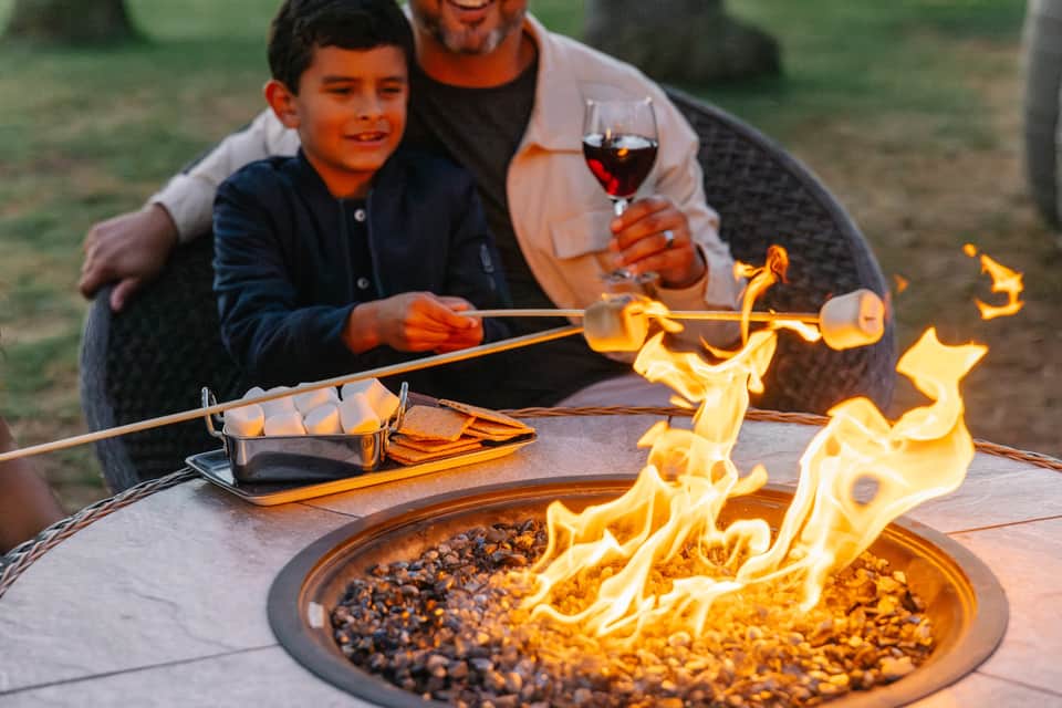 Father and son roasting marshmallows over a fire pit at sunset with wine glass nearby