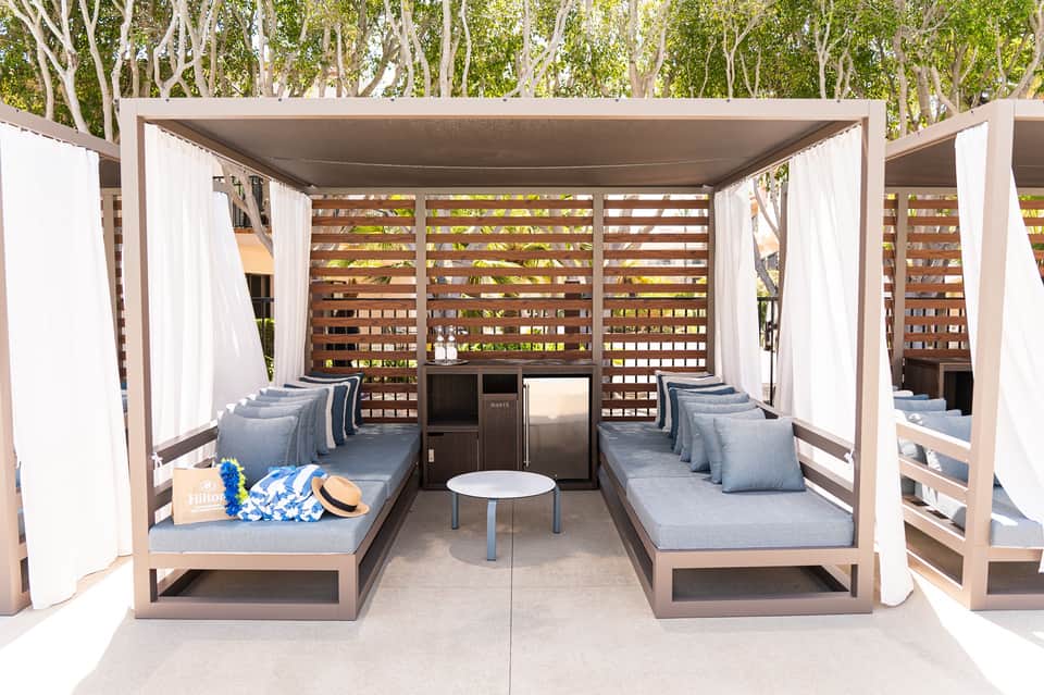 Modern cabana with wooden lattice screens, white curtains, gray cushioned seating, and central table
