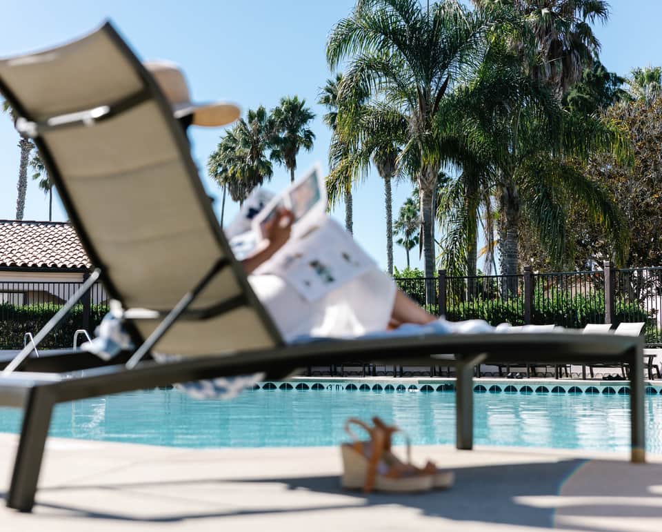 Lounge chair by palm-lined pool with guest relaxing in background under clear sky