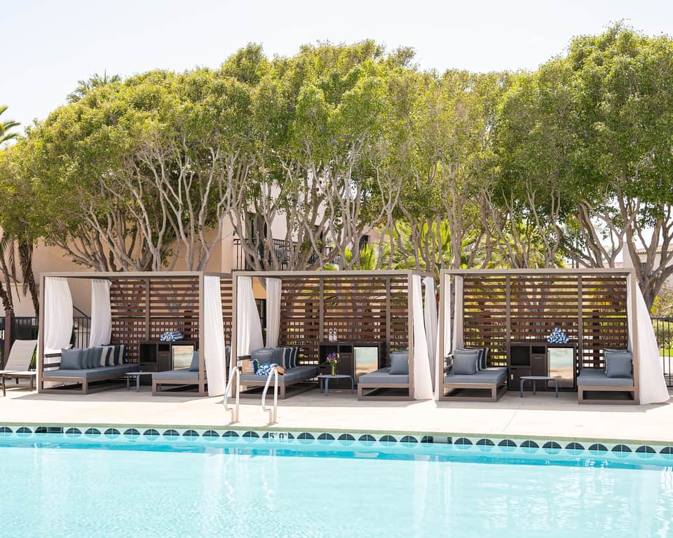 Luxury poolside cabanas with lattice screens, white curtains, and lounge seating overlooking a clear blue pool and tall shade trees