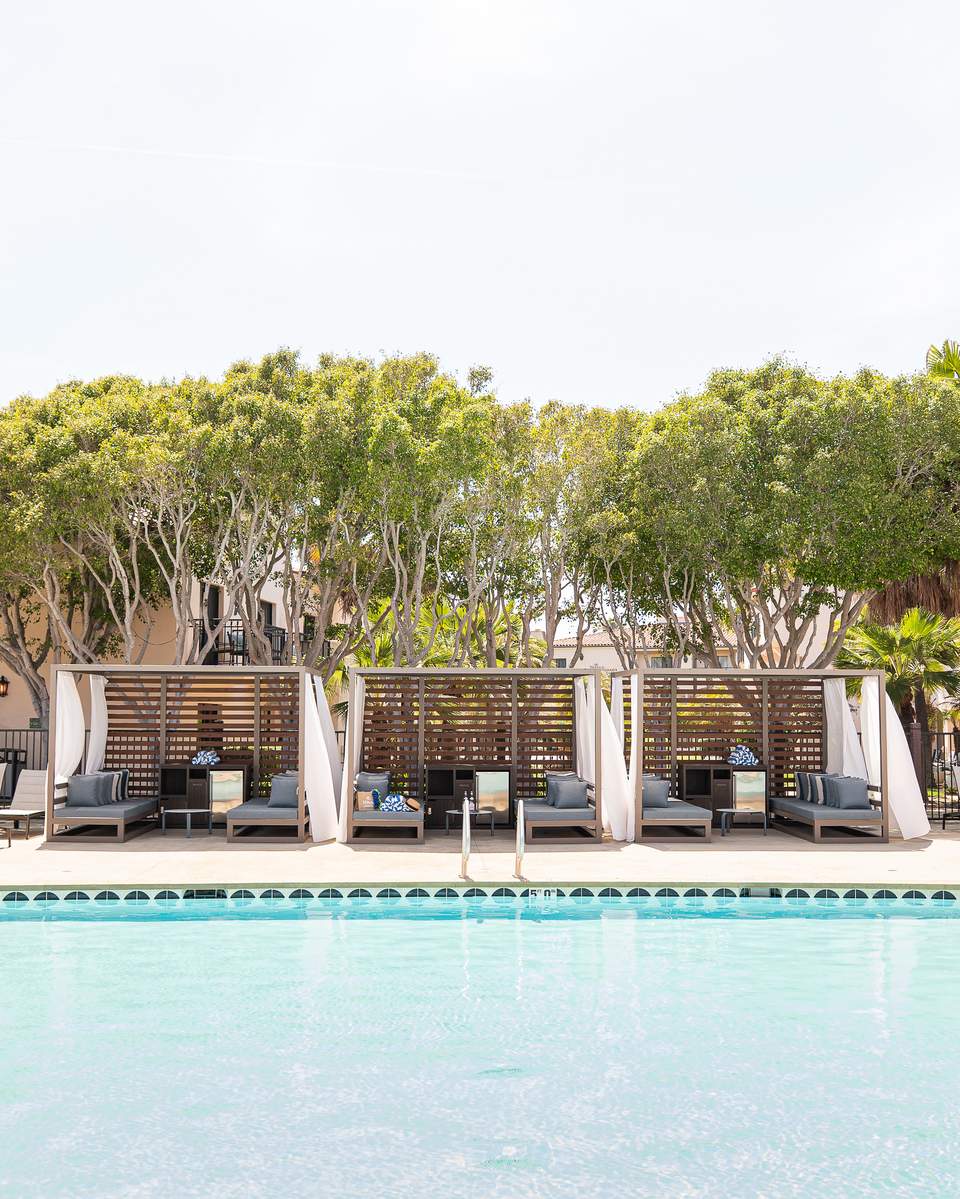 Resort poolside seating area with individual cabanas featuring wooden screens, cushioned lounges, and flowering plants along the pool deck