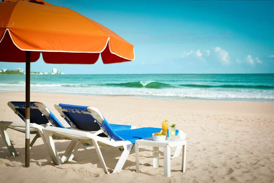 Two lounge chairs with blue cushions under orange umbrella on sandy beach with ocean and blue sky