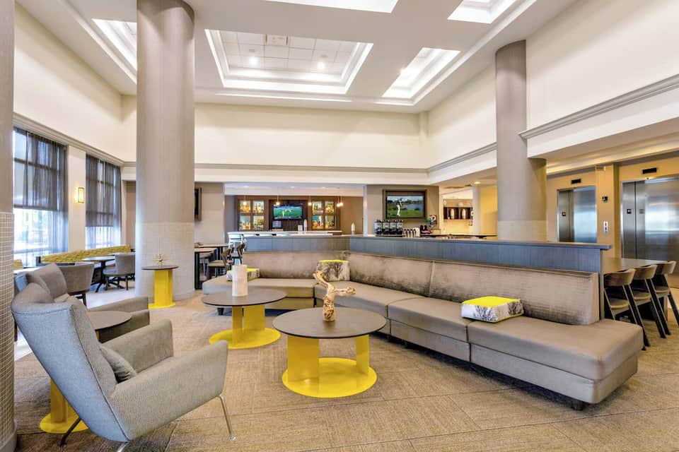Modern hotel lobby lounge with gray seating, yellow accent pillars, skylight ceiling, and service counter