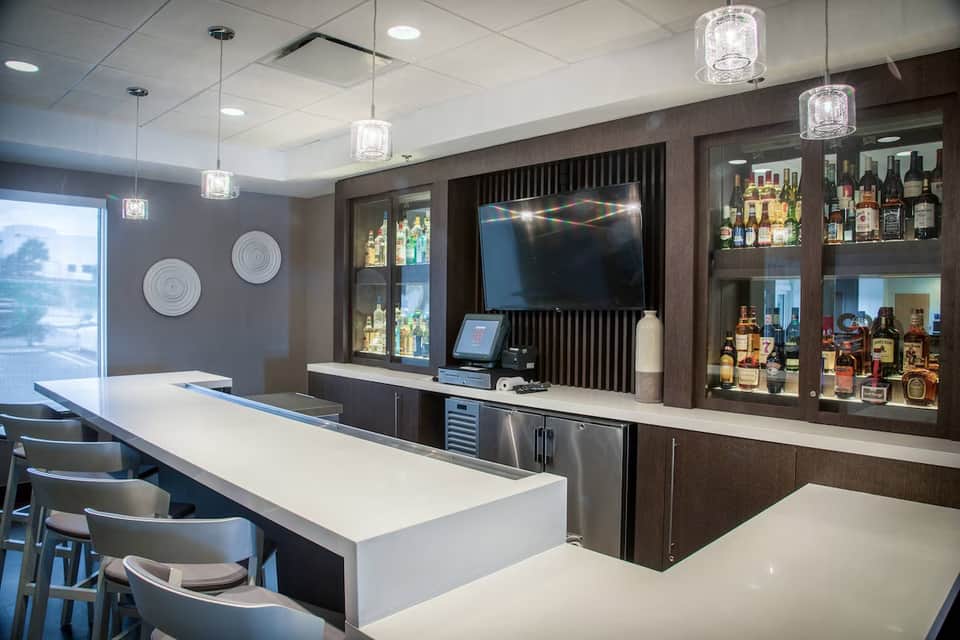 Modern hotel bar with dark wood cabinetry, lit liquor display, and white countertop