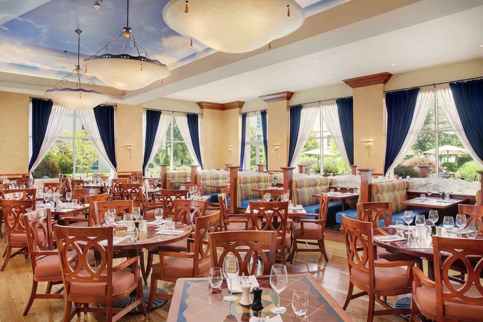 Elegant dining room with round tables, wooden chairs, arched ceiling details, and floor-to-ceiling windows overlooking gardens
