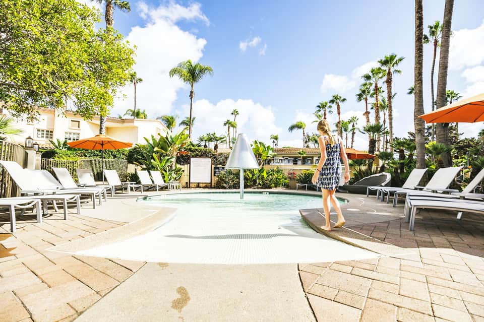 Woman in patterned dress standing by a shallow resort pool with orange umbrellas and palm trees