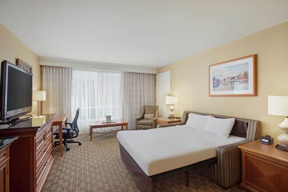 Hotel room with single bed, work desk, flat-screen TV, and seating area overlooking balcony