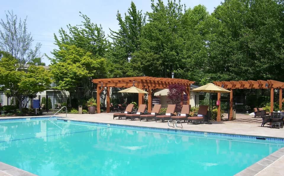 Resort outdoor pool with wooden pergolas, loungers, umbrellas, and lush landscaping on sunny day