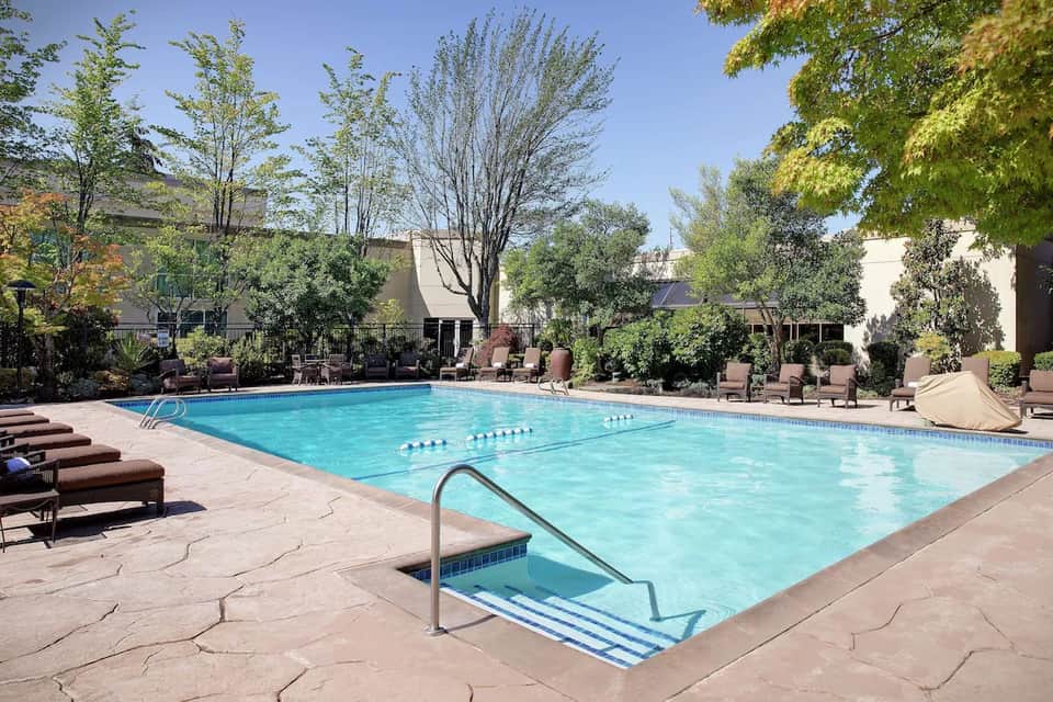 Resort swimming pool with spa steps, lounge chairs, and surrounding hotel buildings with landscaping