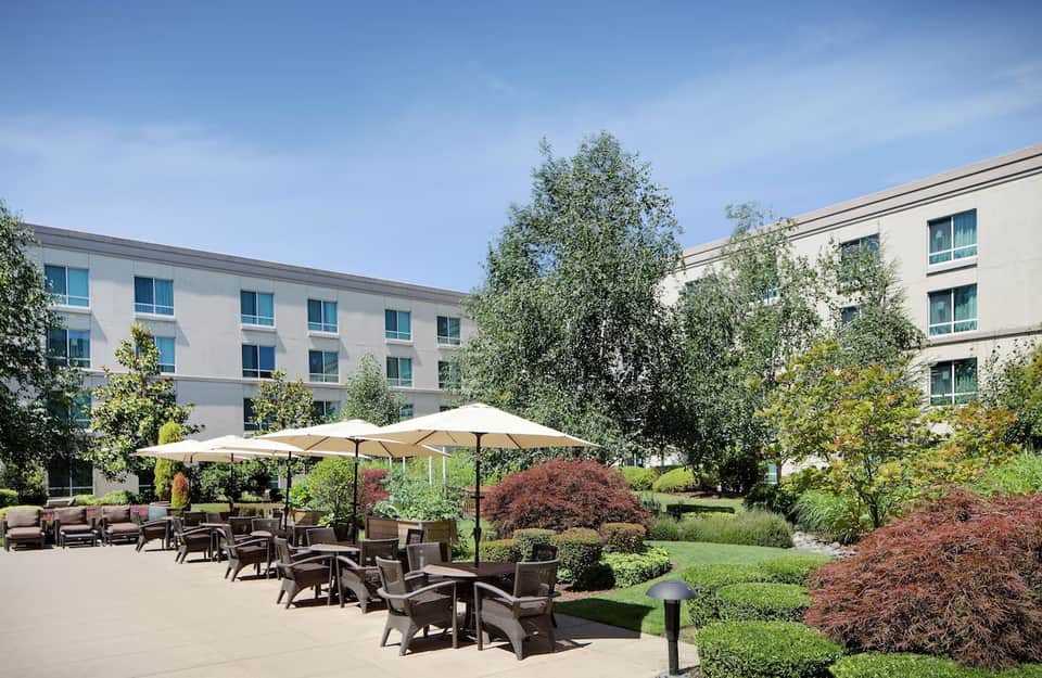 Outdoor courtyard patio with umbrellas, seating, hotel buildings, and manicured gardens under clear sky