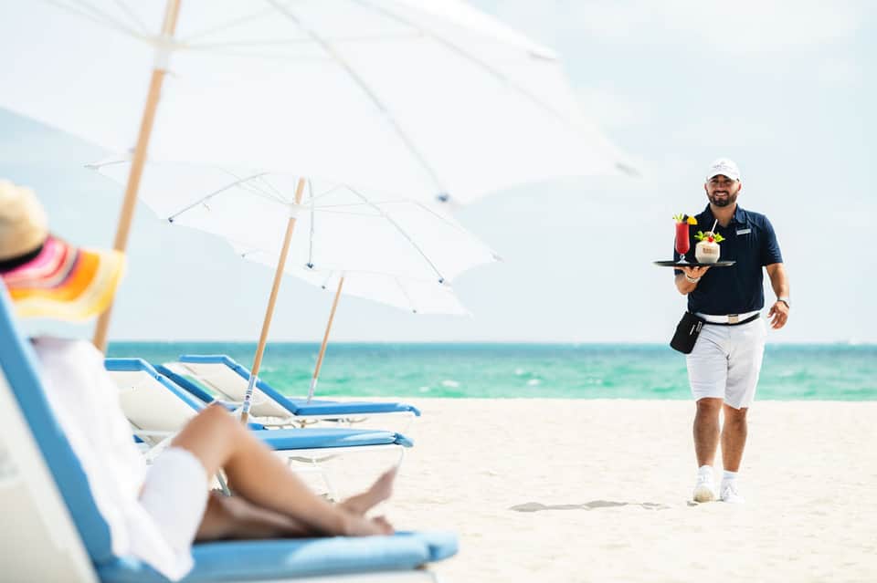 Beach server in black uniform delivering drinks to guests under white umbrellas on sand
