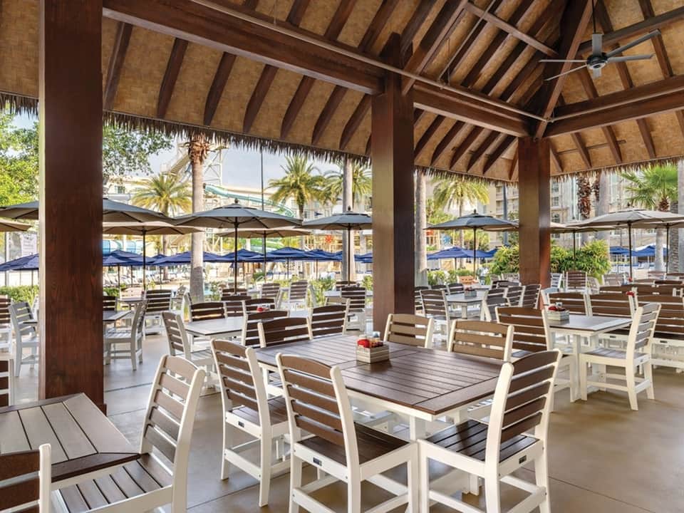 Covered beachfront dining area with wood-slat chairs, umbrellas, palm trees, and ocean views