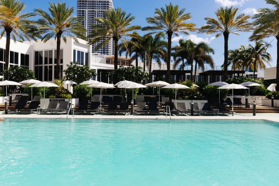 Resort pool area with turquoise water, white umbrellas, palm trees, and modern white buildings