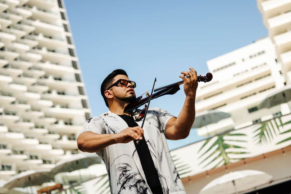 Man playing violin outdoors between modern white hotel buildings against clear sky