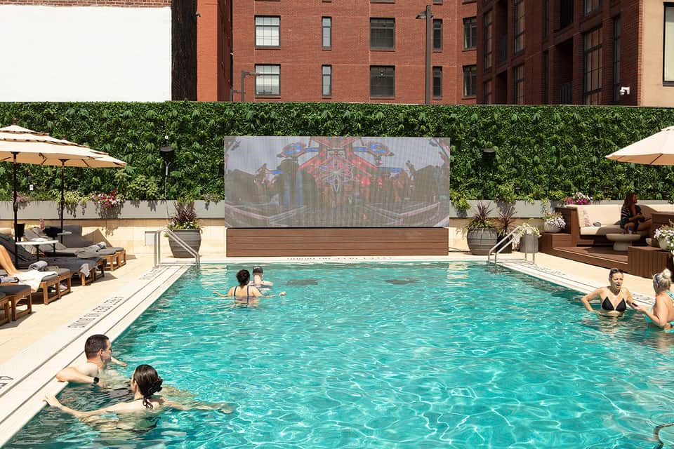 Urban rooftop pool with guests swimming, green hedge walls, brick buildings, and outdoor movie screen