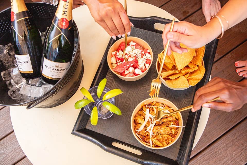 Overhead view of champagne bottles, gourmet appetizers, and lime garnishes on wooden poolside table