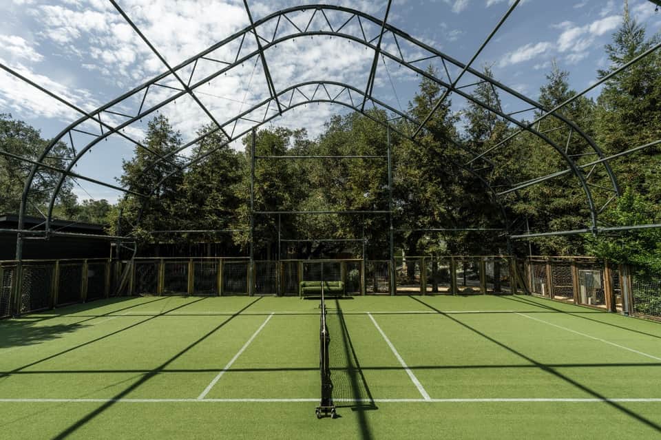 Indoor tennis court with white markings, metal framework roof, and natural lighting