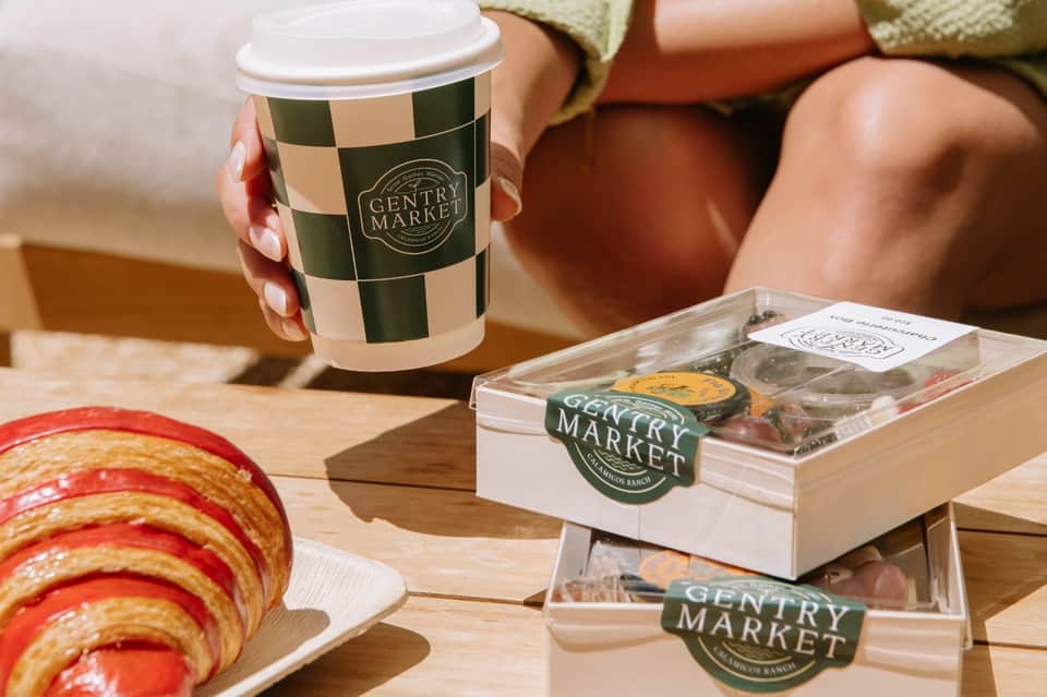 Hand holding checkered coffee cup with Gentry Market pastry box and baked goods on wooden table