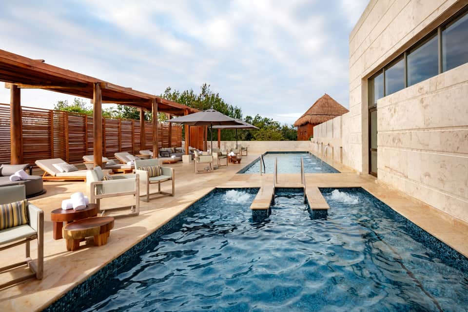 Luxury resort pool deck with lounge chairs, wooden pergola, spa facilities, and stone building with water features