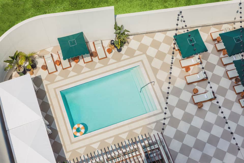 Aerial view of resort pool surrounded by teal umbrellas, wooden lounge chairs, decorative pavers, and white walls