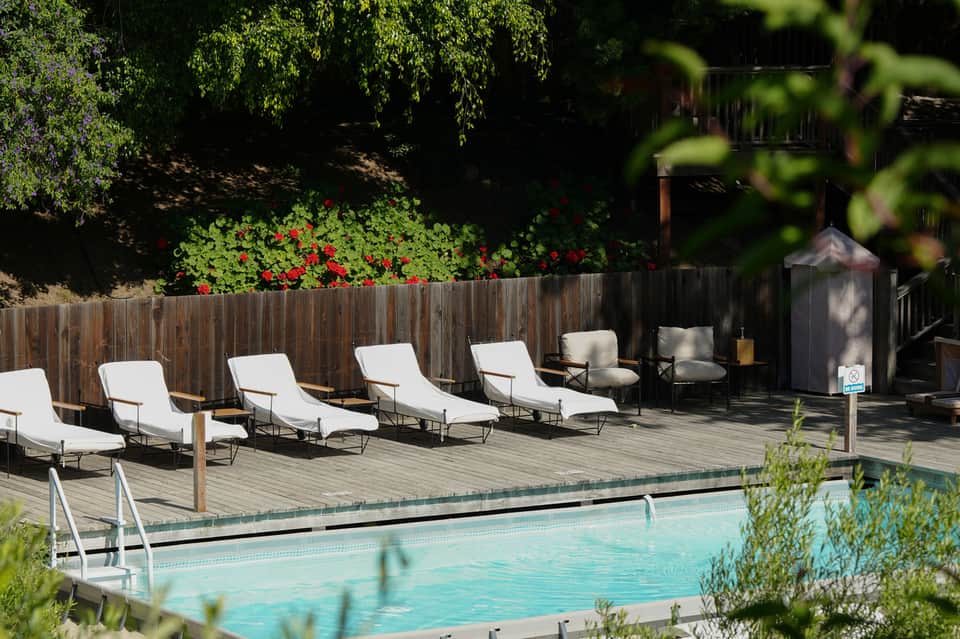 Outdoor pool deck with white lounge chairs, wooden fence, and red flowers under trees