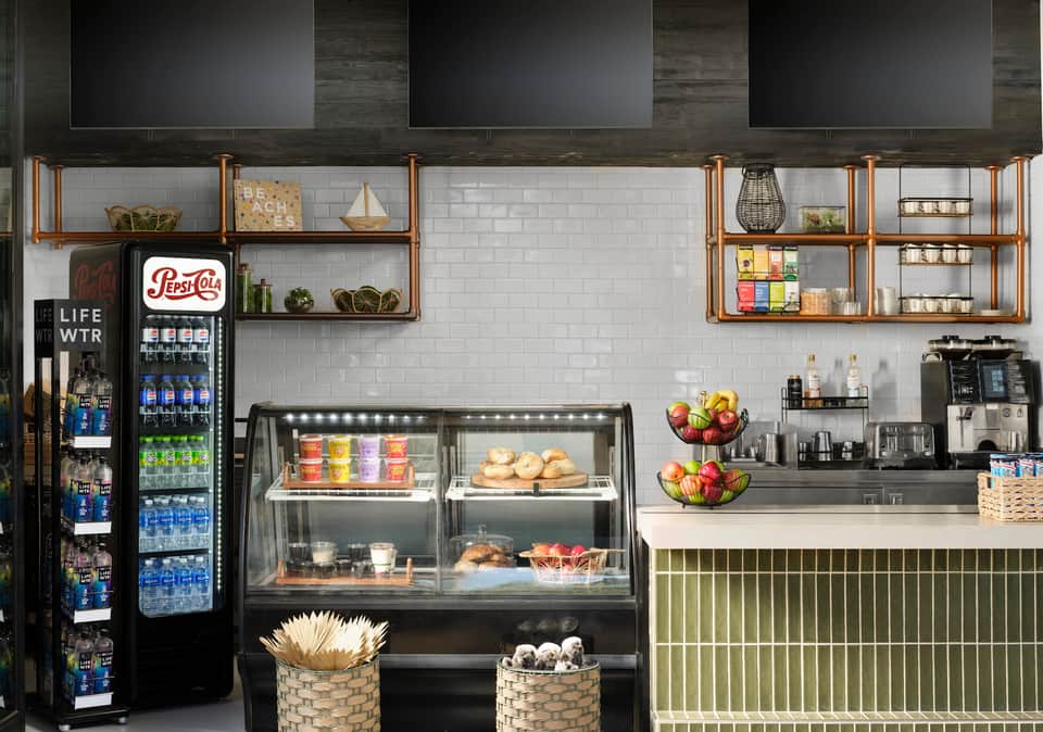Modern cafe counter with white subway tile, wooden shelving, Pepsi cooler, pastry display case, and fresh fruit bowls