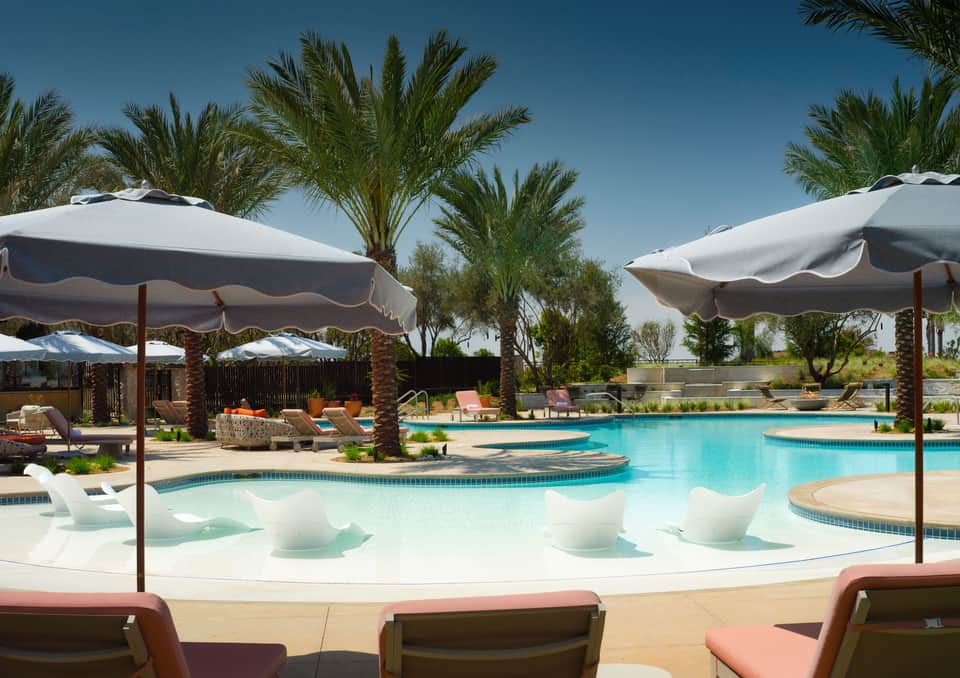 Luxury resort pool with turquoise water, white umbrellas, loungers, palm trees, and shallow spa area under clear blue sky