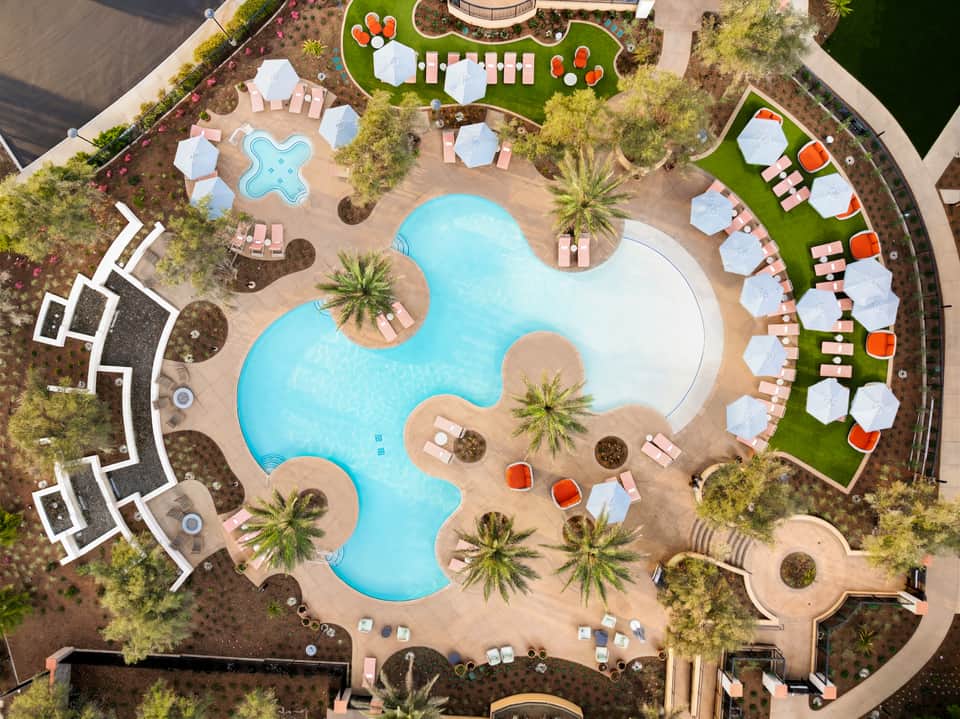 Aerial view of resort pool complex with curved pools, pink lounge chairs, umbrellas, and surrounding palm trees