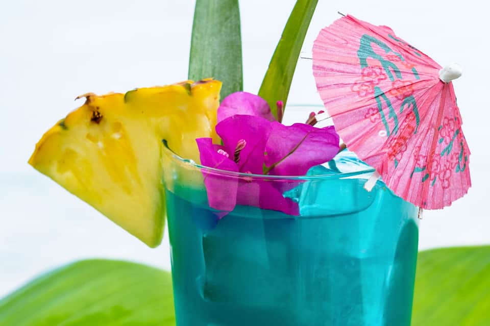 Tropical cocktail with pineapple slice, pink umbrella, magenta flowers in turquoise glass with green straws