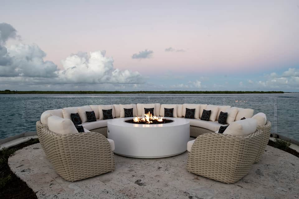 Curved woven lounge seating around fire pit overlooking calm water at dusk
