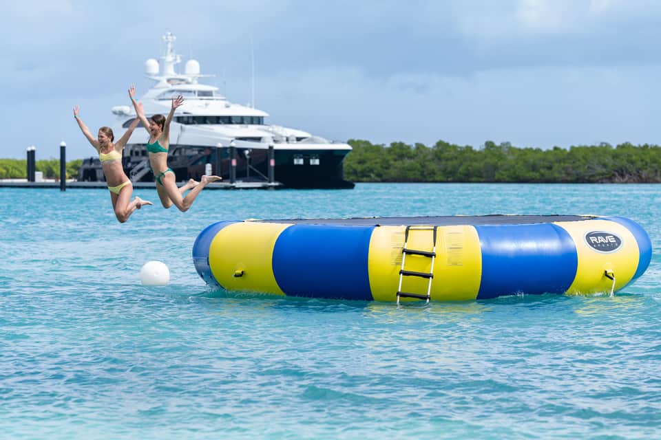 Two children jumping off inflatable water toy with yacht and mangroves in background