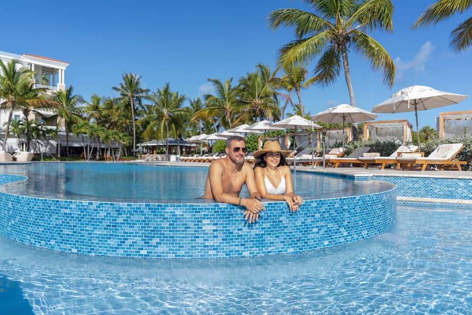 Couple in shallow pool with palm trees, cabanas, and blue sky