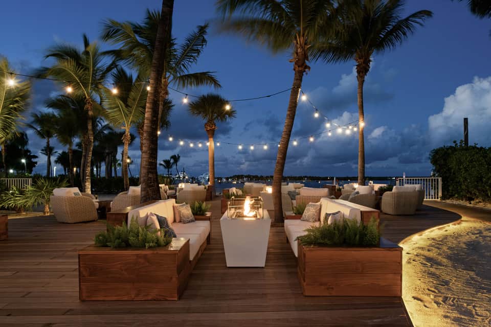 Beachfront lounge area at dusk with fire pit, woven seating, palm trees, and string lights