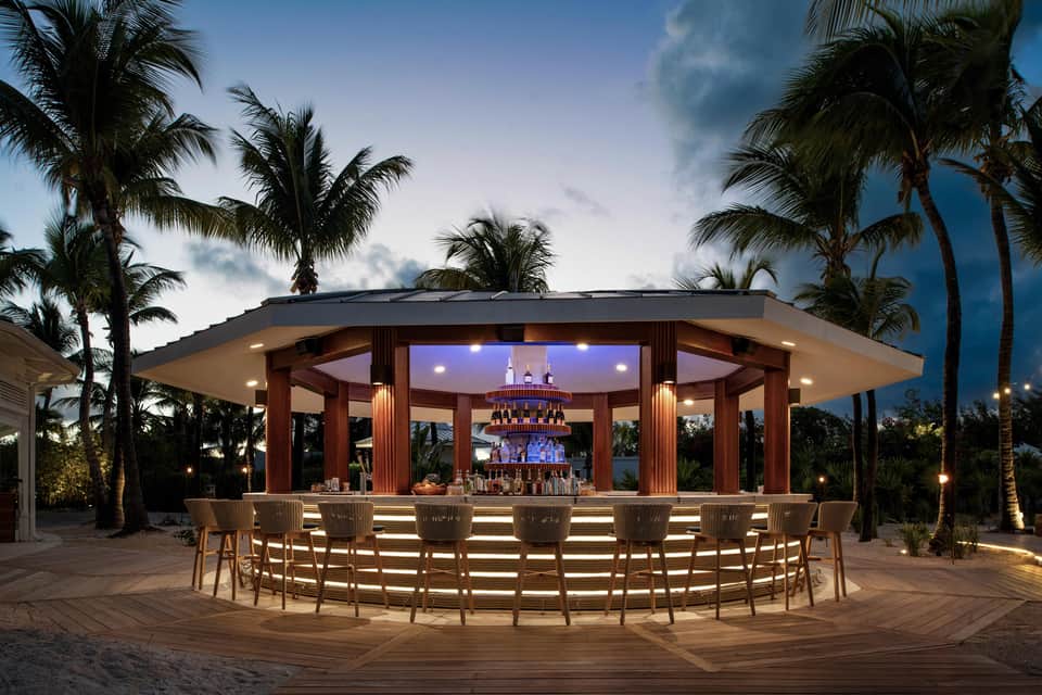 Beachfront tiki bar at dusk with palm trees, illuminated wooden structure, and curved seating for multiple guests