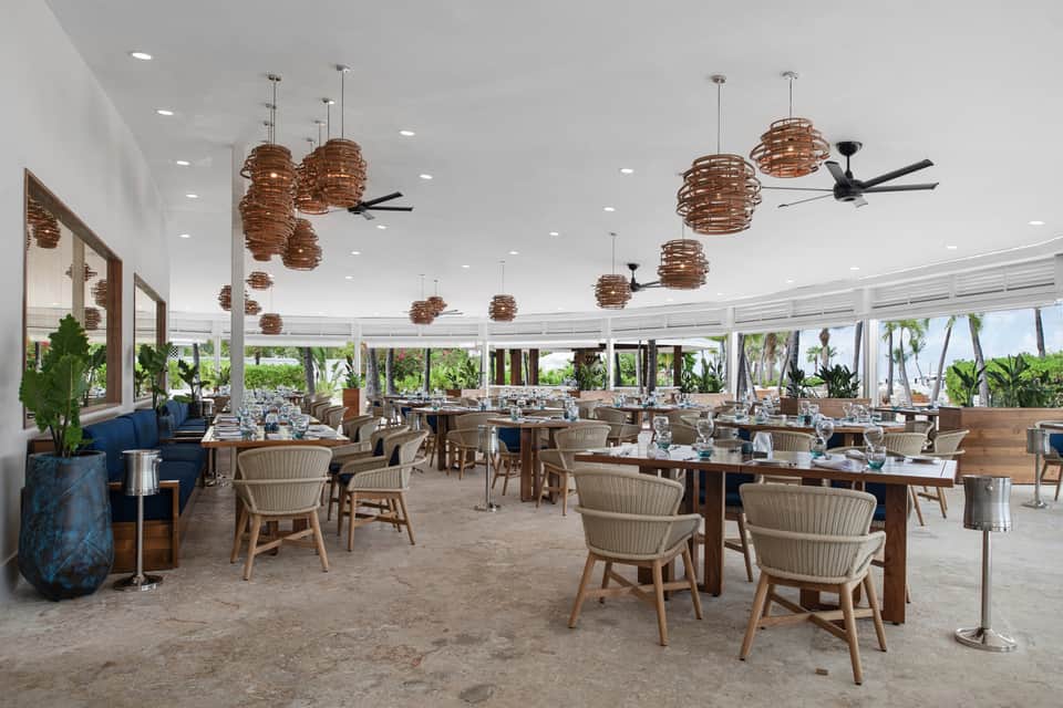 Beachfront restaurant with wooden tables, woven pendant lights, and panoramic ocean views through windows
