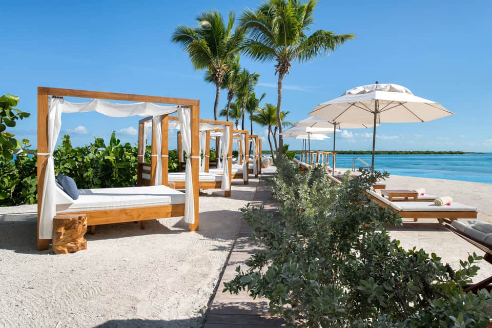 Beachfront cabana beds with white linens and wooden frames, umbrellas, and palm trees along pristine sand