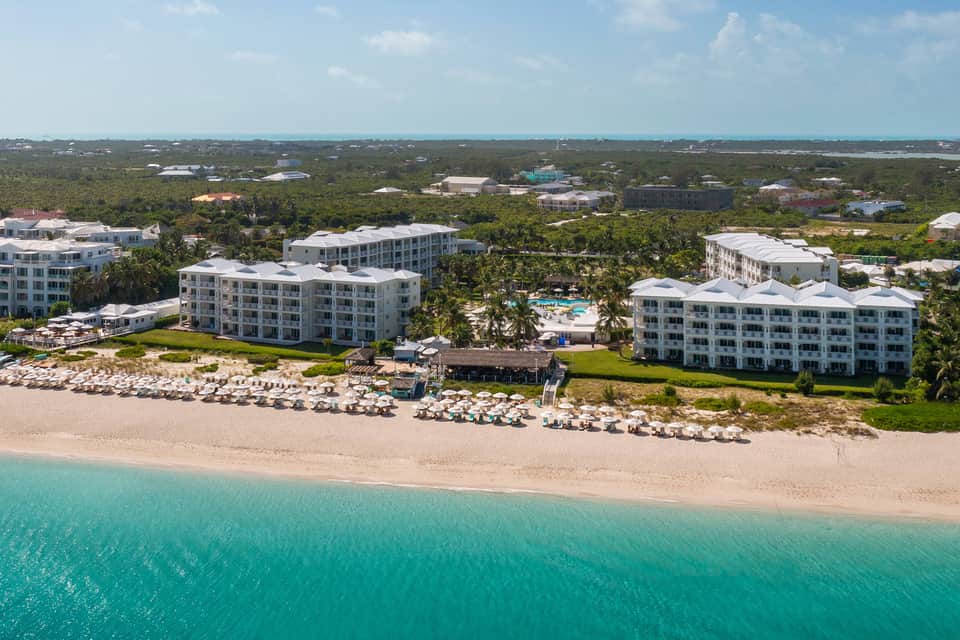 Aerial view of beachfront resort with white buildings, pools, beach loungers, and tropical vegetation