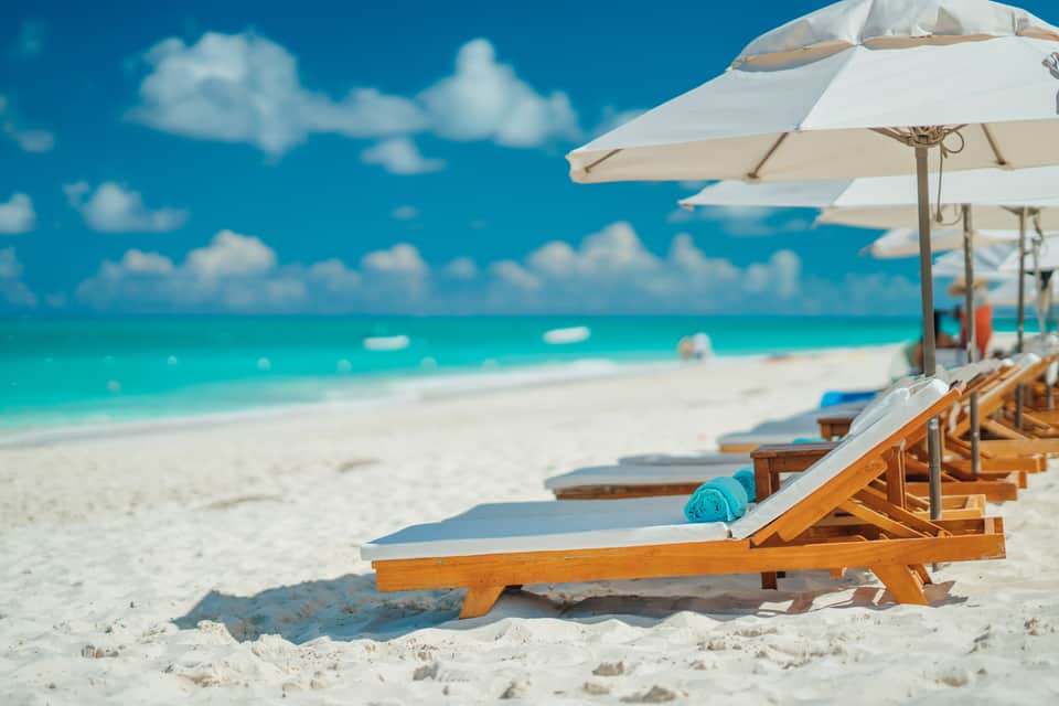 Row of wooden beach loungers with white cushions and umbrellas on white sand with turquoise ocean