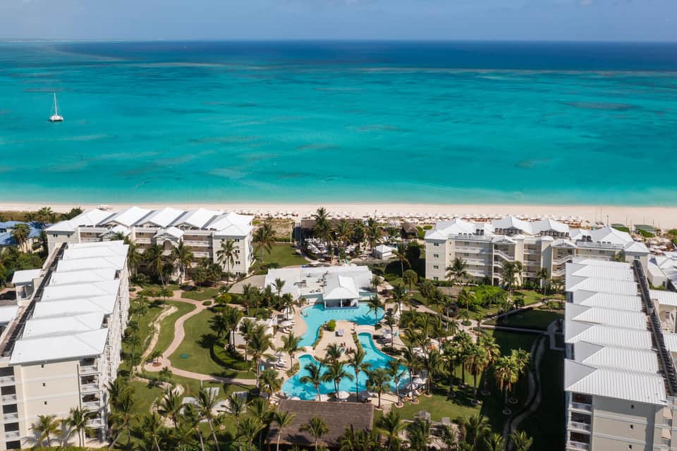 Aerial view of beachfront resort with pools, white buildings, and sailboat on turquoise water