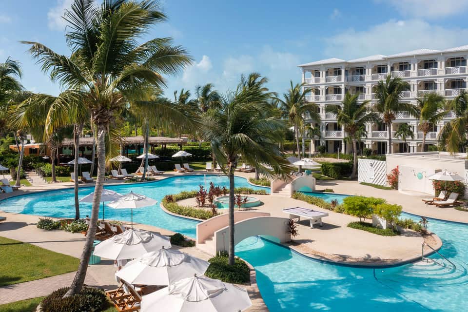 Tropical resort pool area with loungers, umbrellas, palm trees, and white hotel building