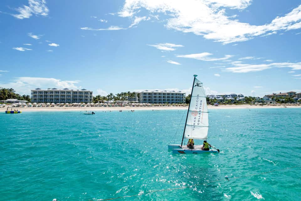 Sailboat with two people on turquoise ocean near sandy beach with resort buildings