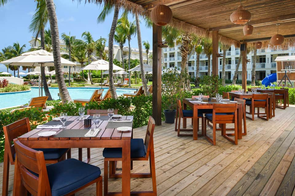 Outdoor dining area with wooden tables overlooking resort pool and palm trees